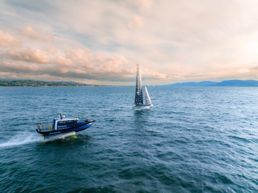 An AC40 America's Cup racing yacht, on Belfast Lough on a warm evening.  It sails over gentle blue waves next to an electic foiling boat.